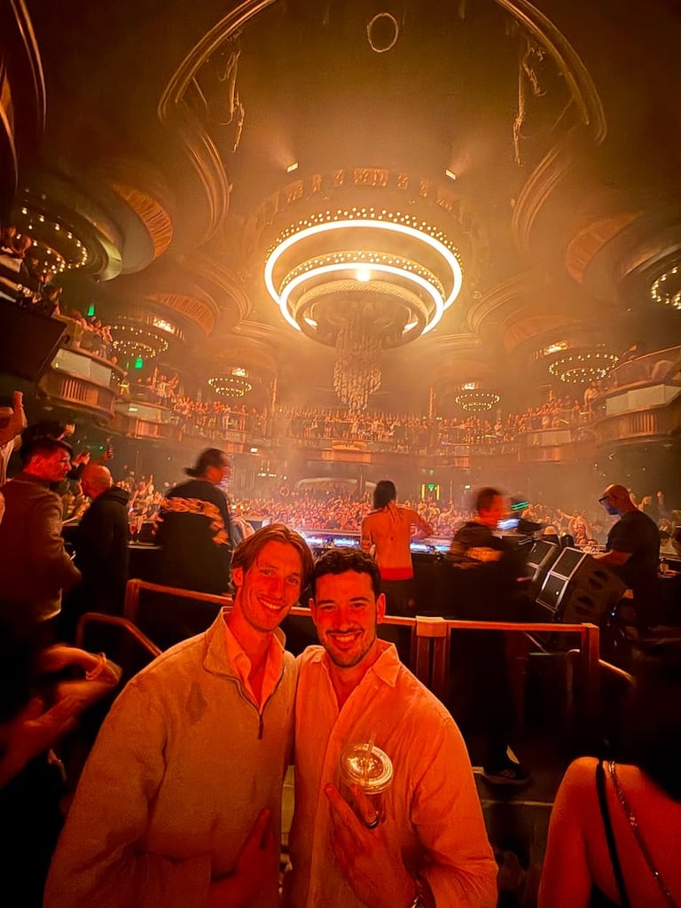 Two men smiling at camera inside ornate golden theater with glowing ceiling dome and crowd in background