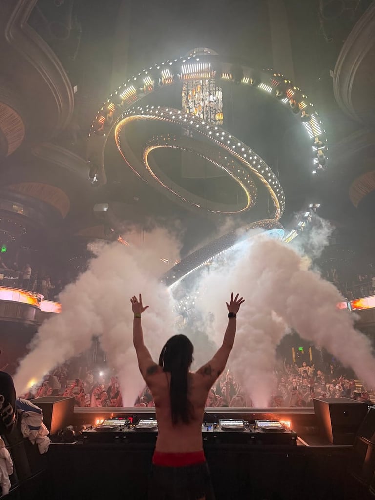 Person with raised arms at concert stage surrounded by white smoke and elaborate illuminated set design overhead